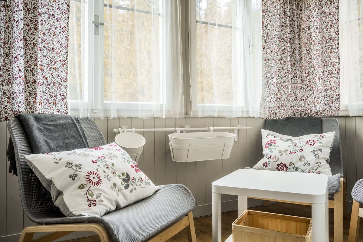 A cozy room with a grey sofa, a small white table, and a wooden bench. There are two white side tables and a white basket on the wall. The room has floral curtains and a view of trees through the window.