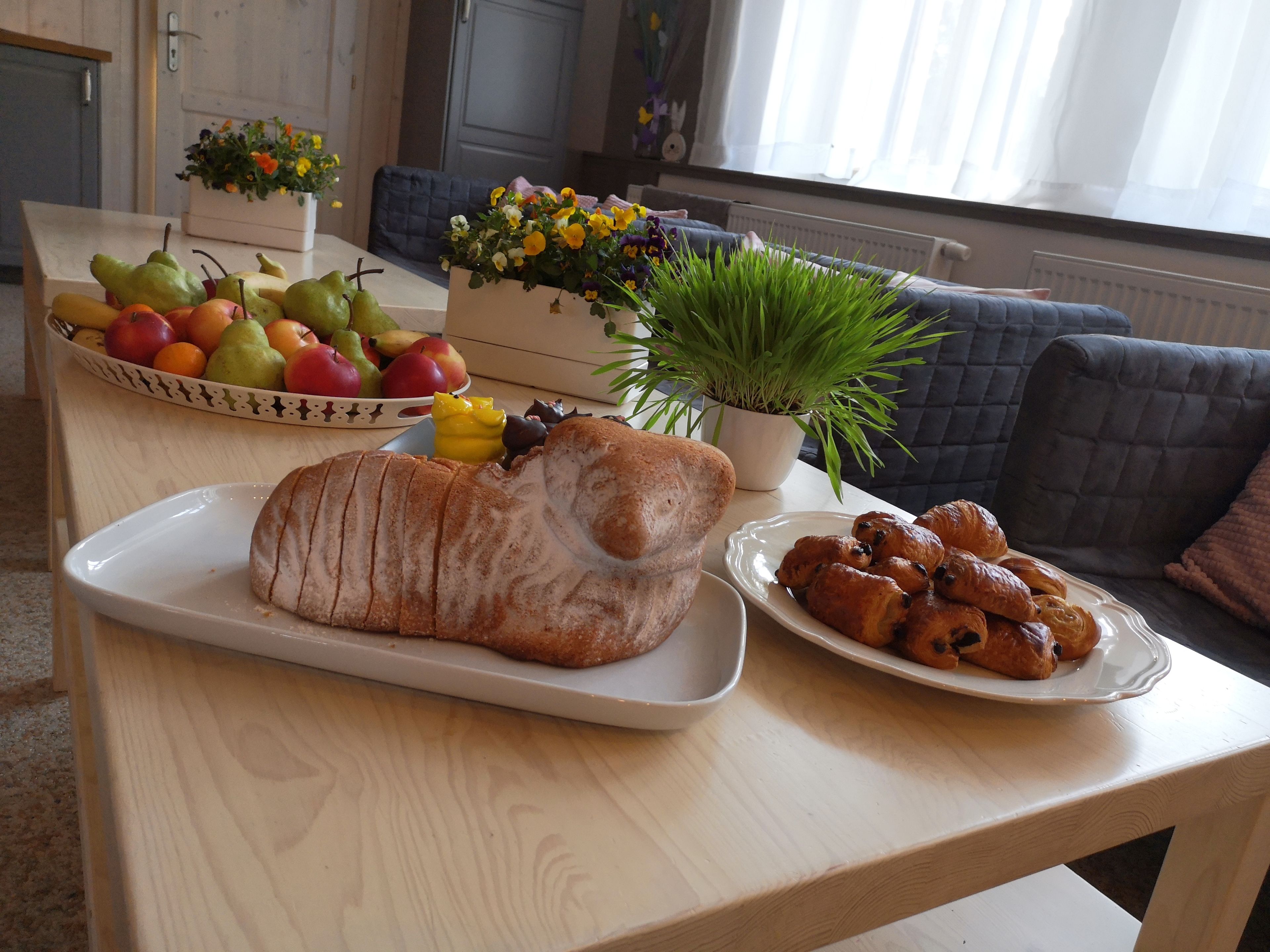 A dining table with a loaf of bread, a plate of pastries, a bowl of fruit, and a potted plant.