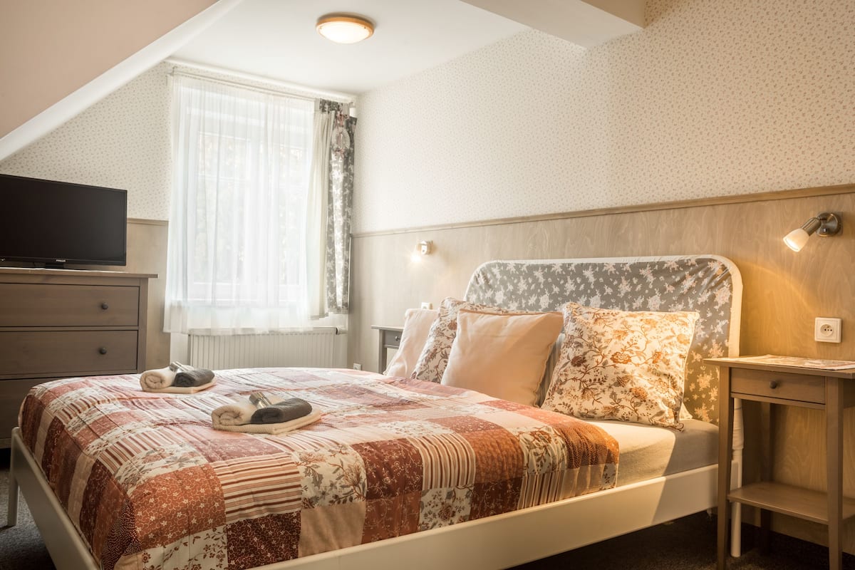 A bedroom with a bed, a television on a dresser, a window with curtains, and a nightstand with a lamp.