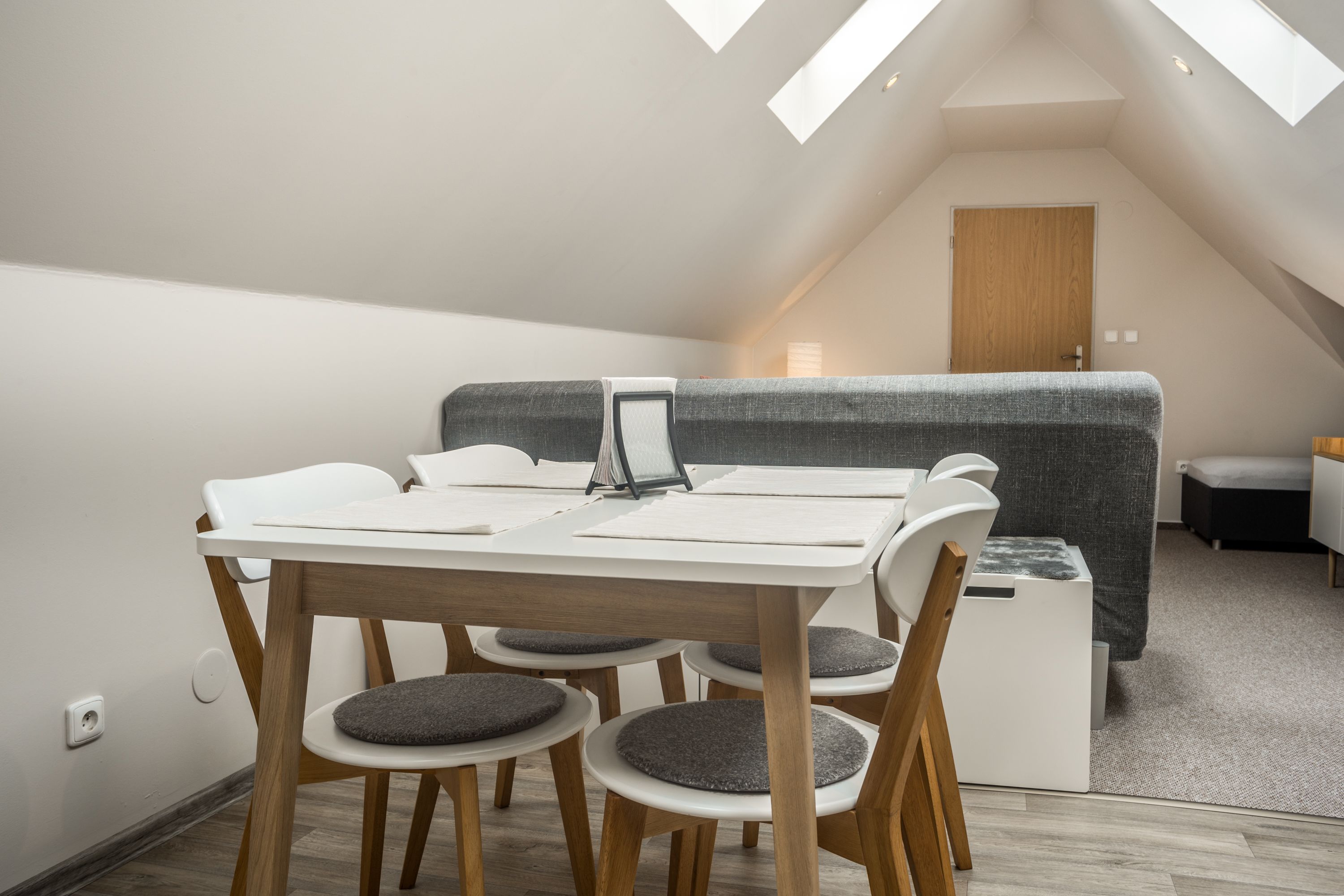 Un dormitorio estilo loft moderno con techo inclinado, una mesa de comedor de madera con sillas, un sofá gris y un pequeño armario blanco.