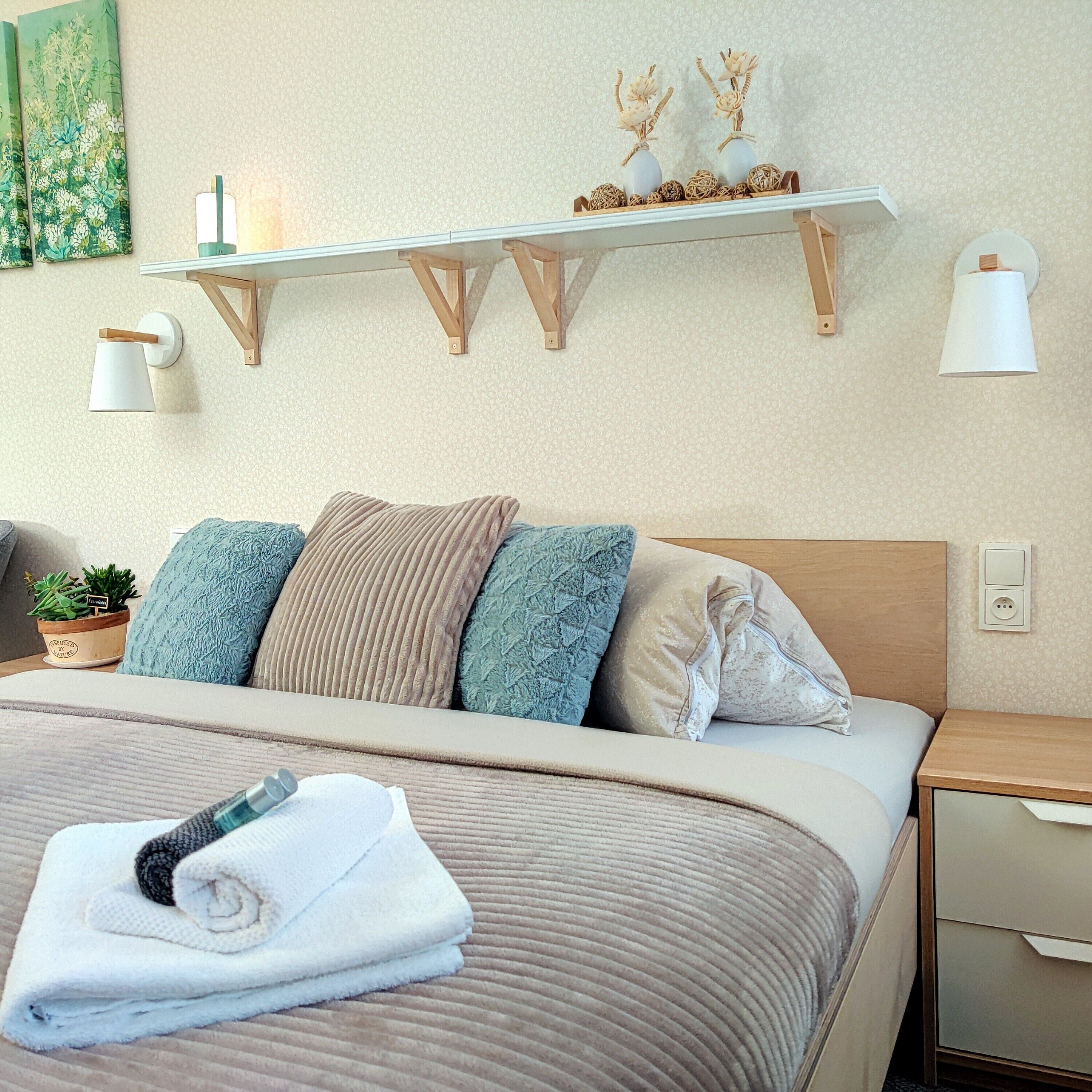 A neatly made bed with a beige comforter and multiple pillows, a bedside table with a lamp, a small plant, and a wall-mounted shelf with decorative items.