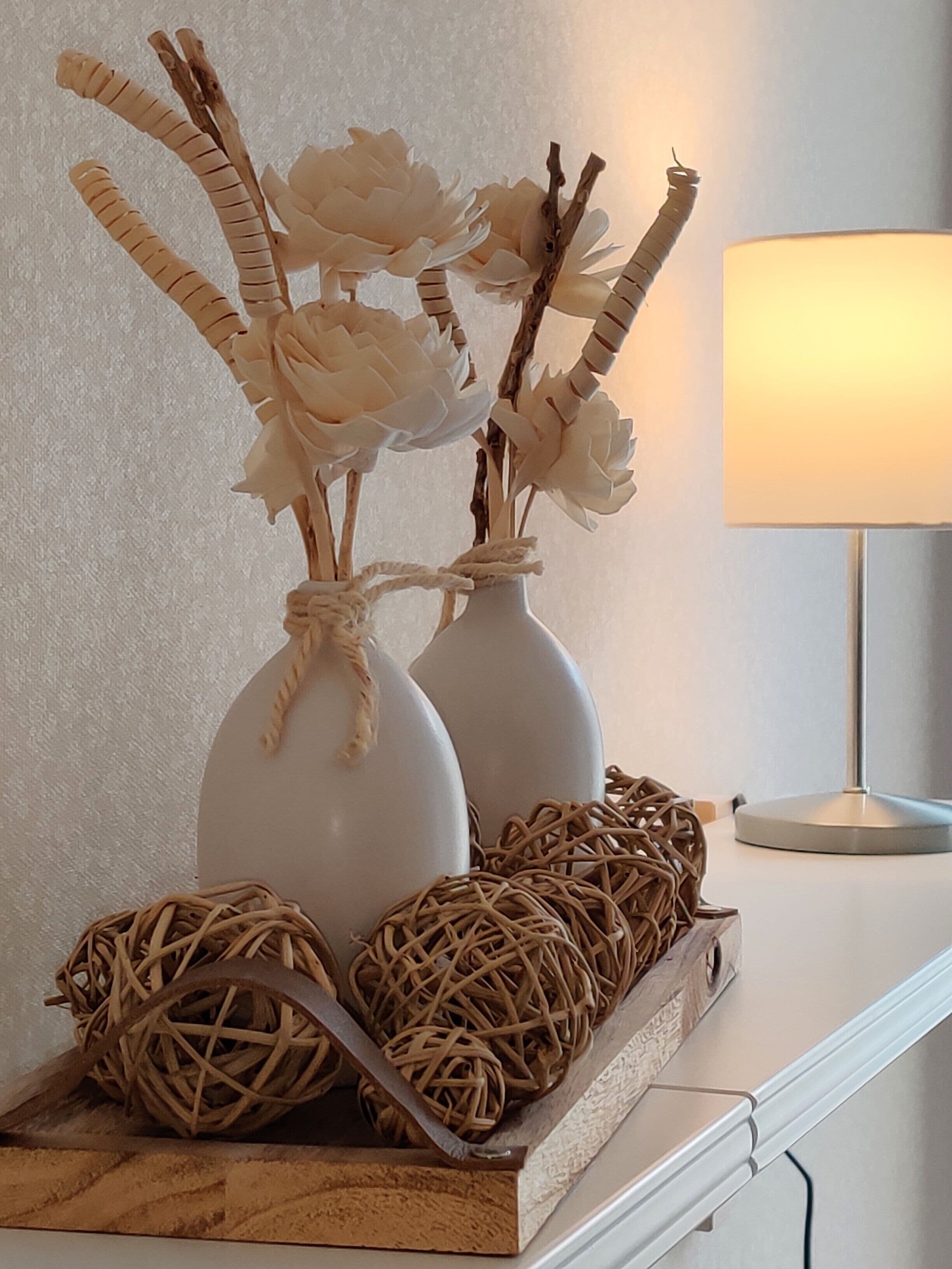 Two white vases with paper flowers and wicker balls on a shelf.