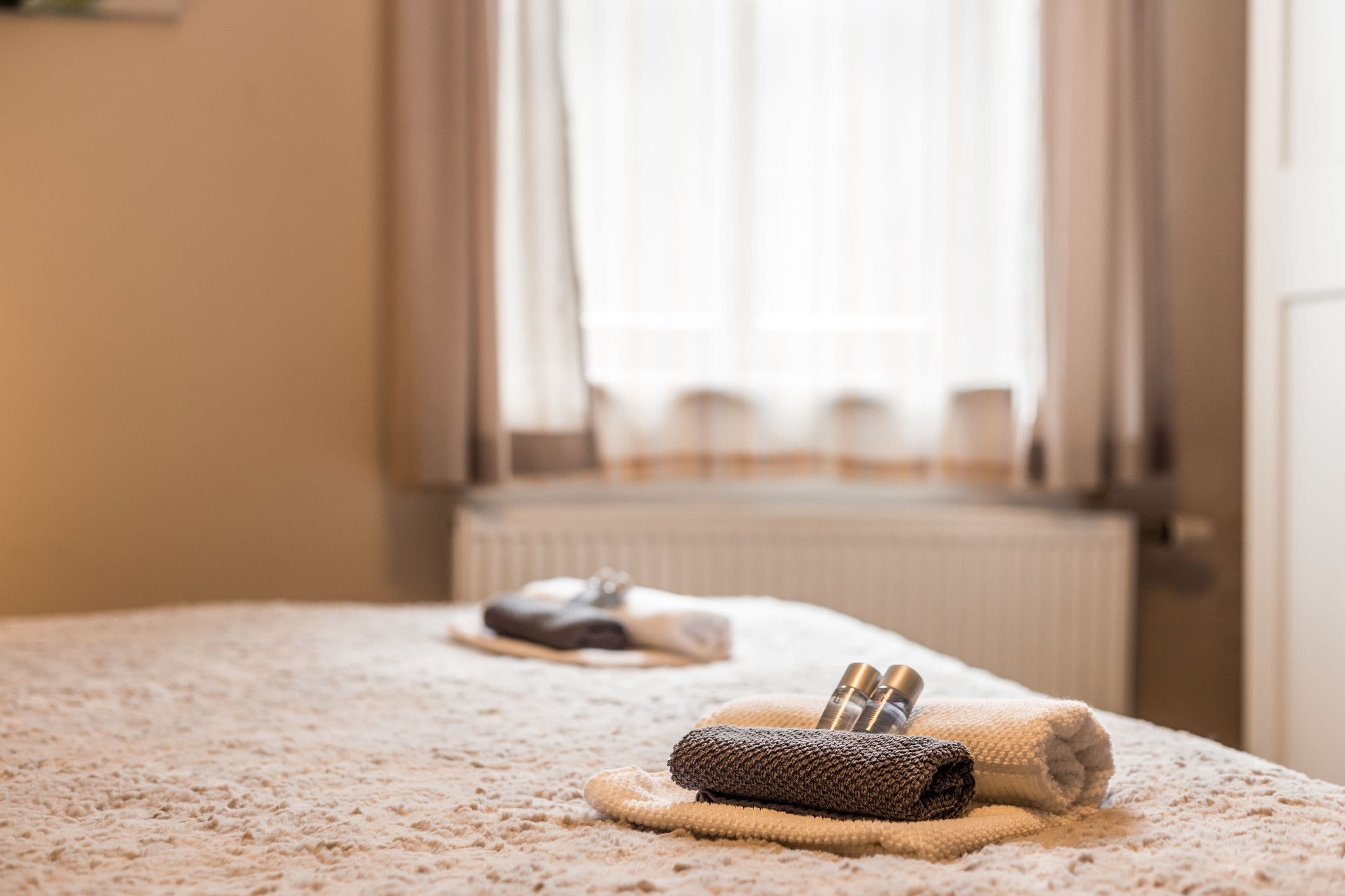 Ein ordentlich bezogenes Bett mit einem zusammengerollten Handtuch und Hausschuhen, einem Heizkörper und einem fenster mit Vorhängen.
