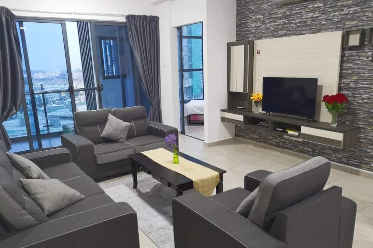 A modern living room with grey sofas, a coffee table, and a flat-screen TV. There is a stone accent wall and a large window with curtains.