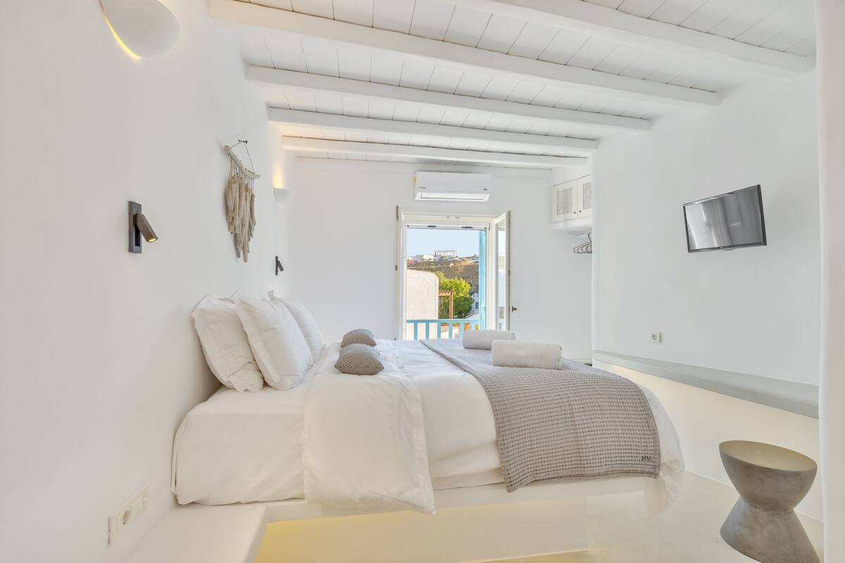 A minimalist bedroom with a bed, a bedside table, a wall-mounted TV, and a view of the outdoors.