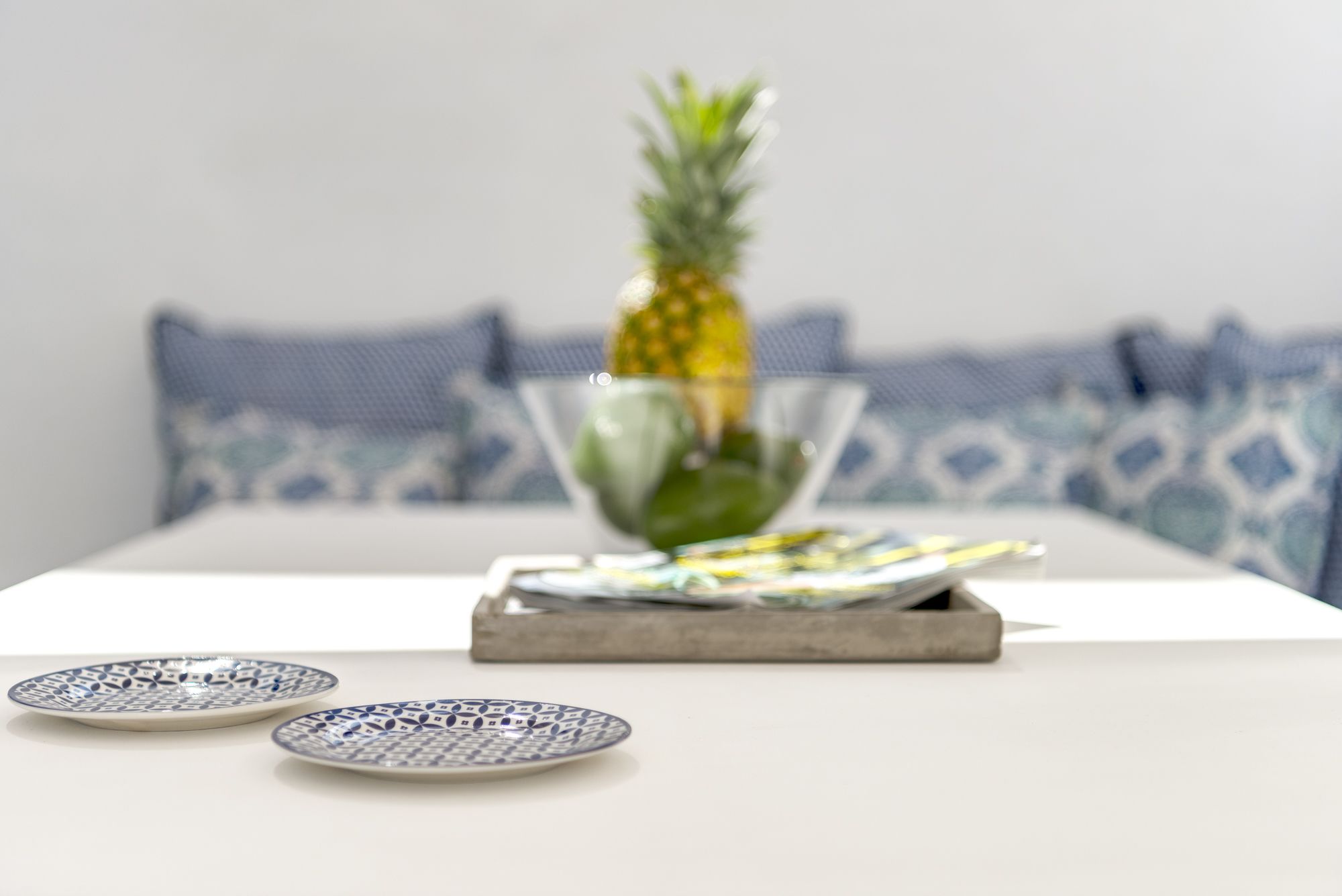 Une table à manger avec un ananas et un saladier de fruits, deux assiettes à motifs et un plateau sur une surface blanche, avec des coussins à motifs bleus en arrière-plan.