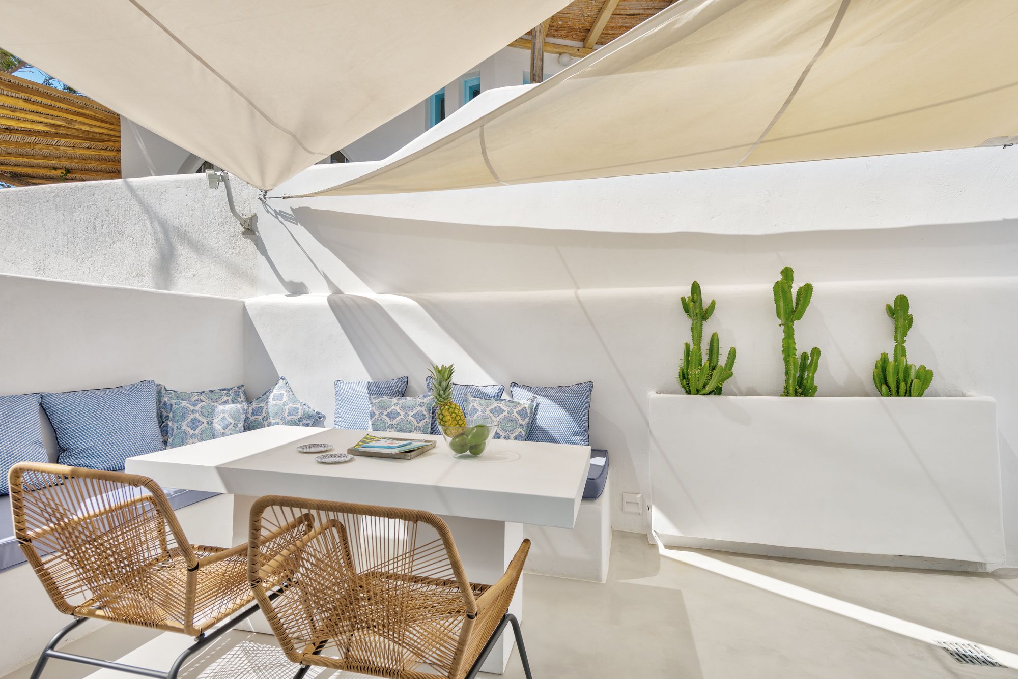Un área de estar exterior moderna con una mesa blanca y sillas de mimbre, un banco blanco acolchado y tres cactus en macetas.