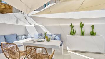 Un área de estar exterior moderna con una mesa blanca y sillas de mimbre, un banco blanco acolchado y tres cactus en macetas.