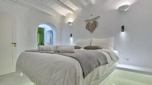 A modern bedroom with a large bed, a hanging light fixture, and a view of the bathroom. - Mykonian Mews (Mykonos)