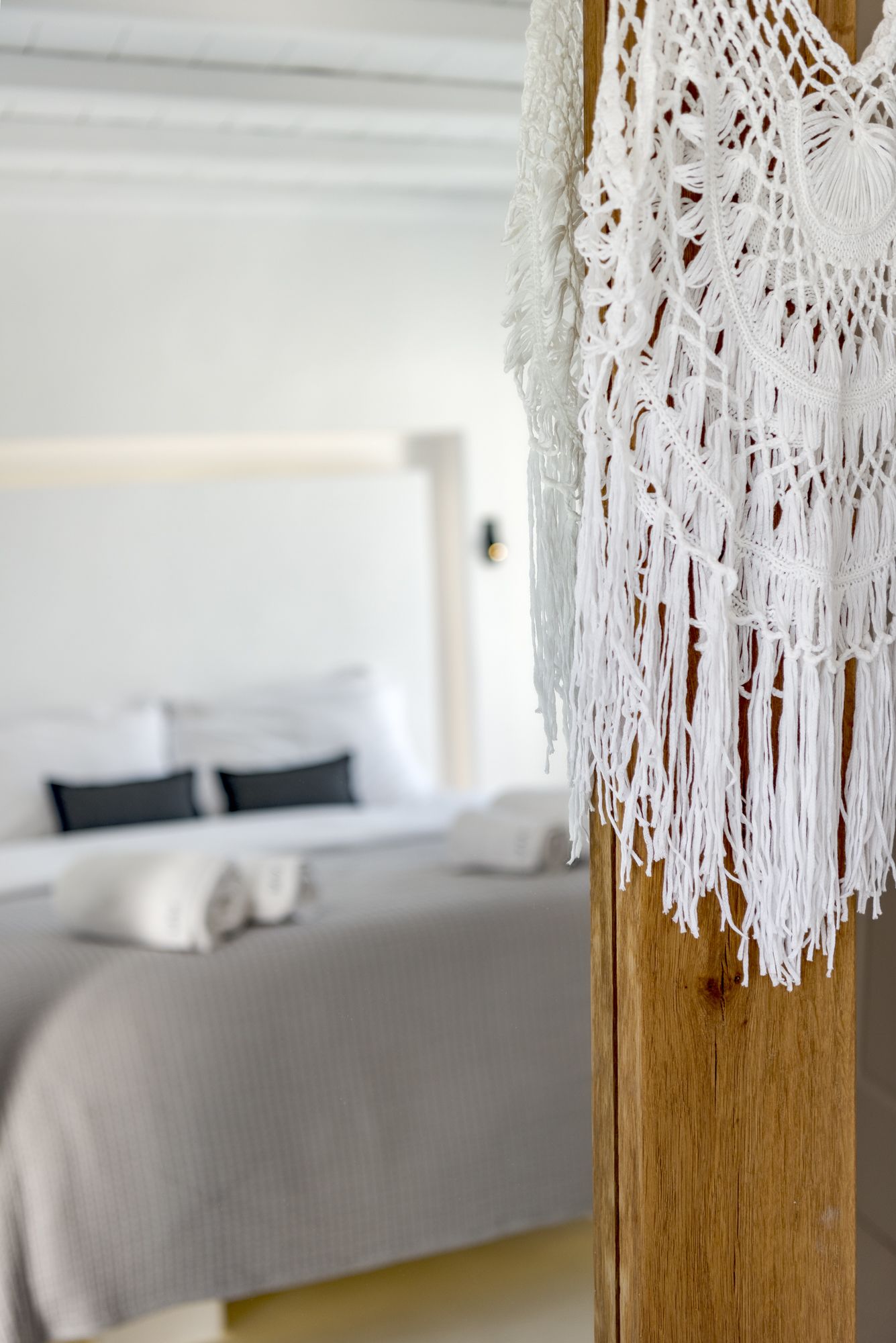Ein Bett mit weißem Makramee-Kopfstück in einem Raum mit Holzkopfstück und grauer Bettdecke.