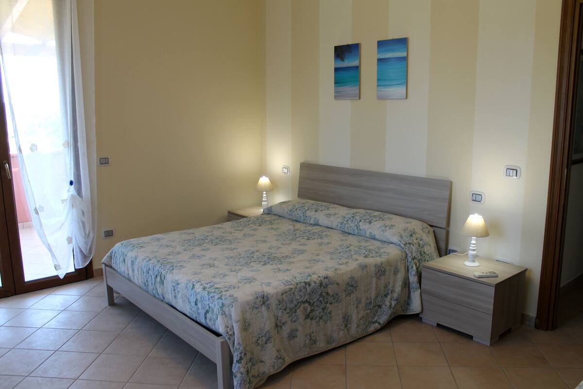 A bedroom with a bed, two bedside tables with lamps, a door, and two wall-mounted paintings.