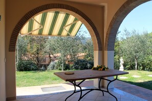 Un patio con tavolo e sedie, circondato da un muro in mattoni e affaccio su un giardino con alberi e una statua.