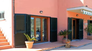 A building with a pink facade, green shutters, and a tiled entrance. There are potted plants on the steps. - Appartamenti La Risacca (Capoliveri)
