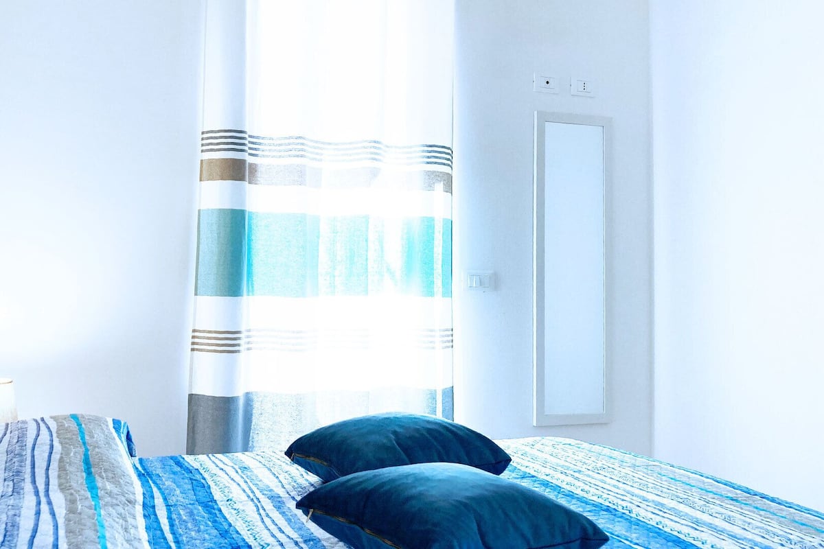 A bedroom with a bed, pillows, a bedside table, a lamp, a window with curtains, and a door.