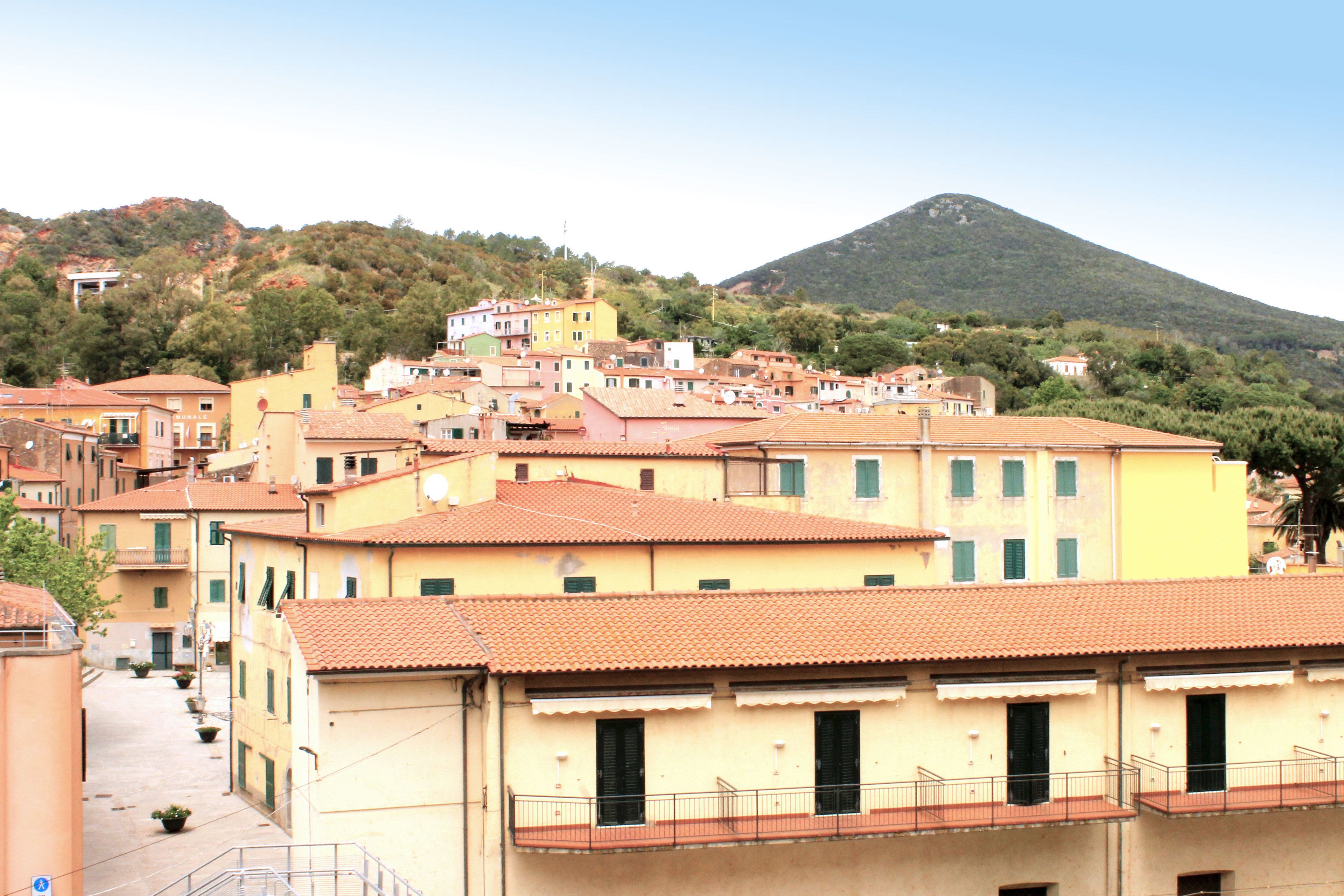 A hillside town with terracotta-roofed buildings, a prominent hill in the background, and a clear sky.