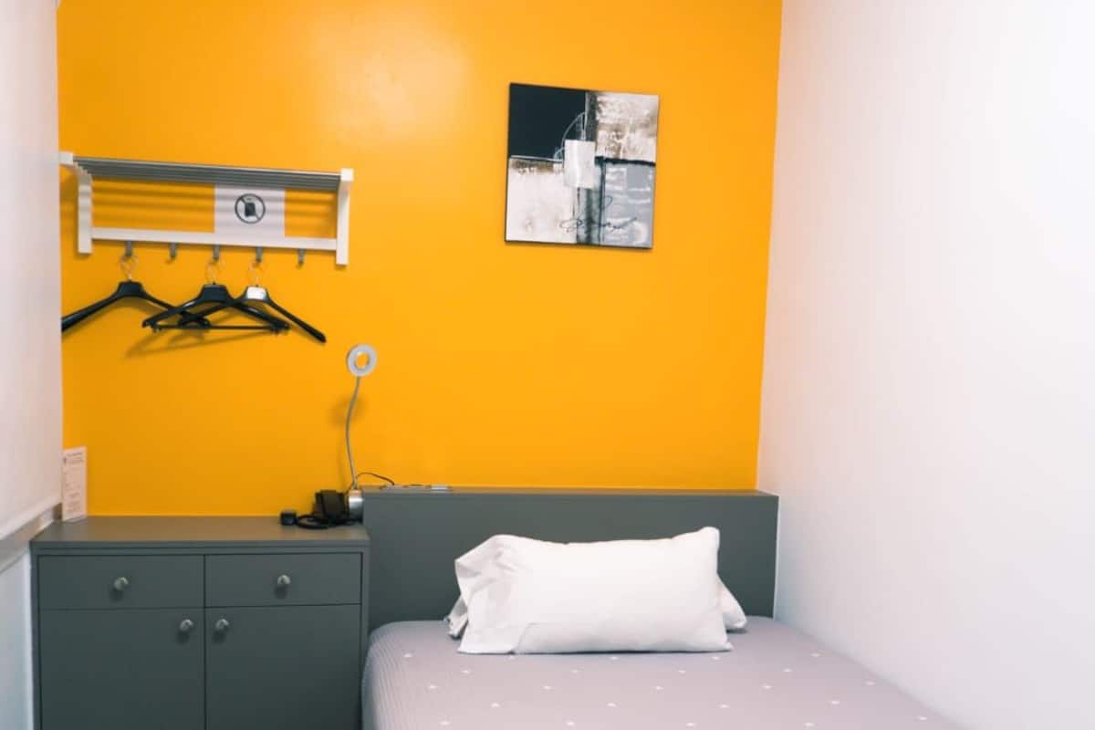A bedroom with a bed, a grey wardrobe, a yellow wall, and a painting.