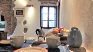 A dining area with a rustic brick wall, a window with blinds, a coffee maker, and a bowl of fruit on the table. - Da Gabriella e David (Marciana)