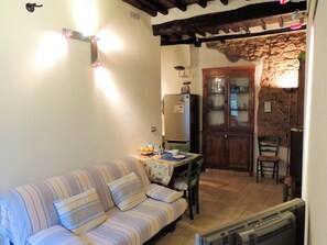 A cozy living room with a striped sofa, a dining table with a bowl of fruit, a refrigerator, and a stone wall.