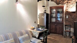 A cozy living room with a striped sofa, a dining table with a fruit bowl, and a kitchen area with a refrigerator and wooden cabinets. - Da Gabriella e David (Marciana)