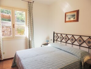 A bedroom with a bed, a window with curtains, a bedside table with a lamp, and a framed picture on the wall.