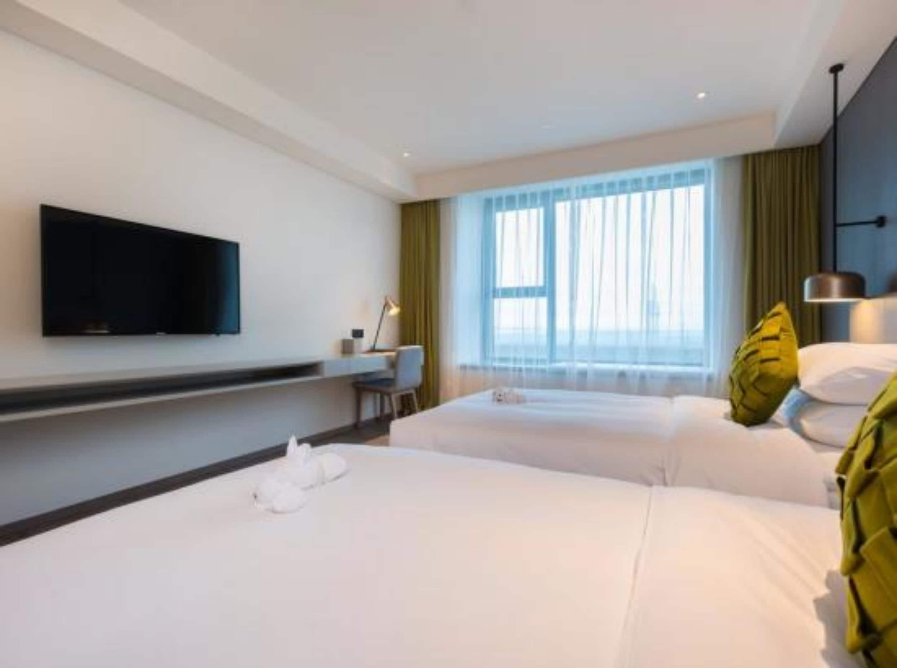 Habitación de hotel con dos camas, un escritorio, un televisor de pantalla plana en la pared y un ventanal con cortinas traslúcidas.