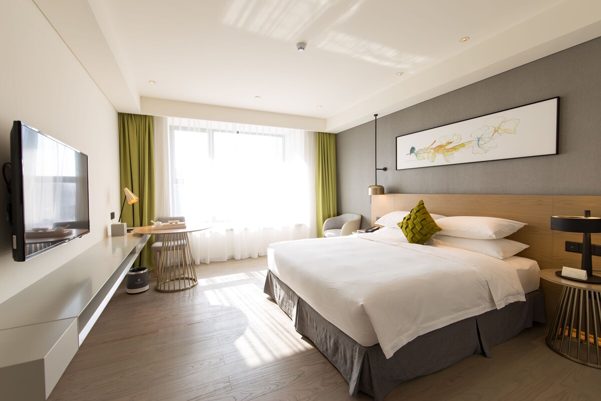 Une chambre d’hôtel moderne dotée d’un grand lit, d’un téléviseur à écran plat, d’un bureau et d’une grande fenêtre avec des rideaux.
