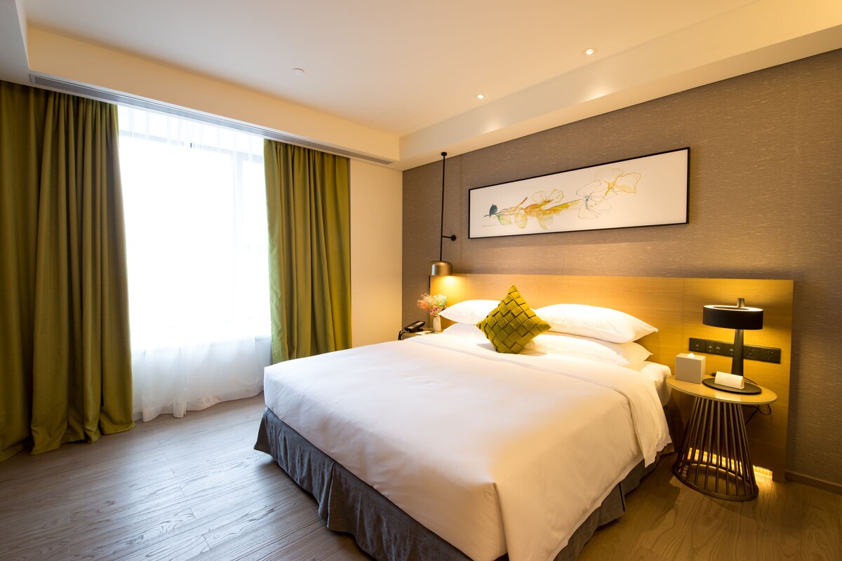 Une chambre d’hôtel avec un grand lit, une table de chevet avec une lampe, une œuvre d’art accrochée au mur et une fenêtre avec des rideaux.