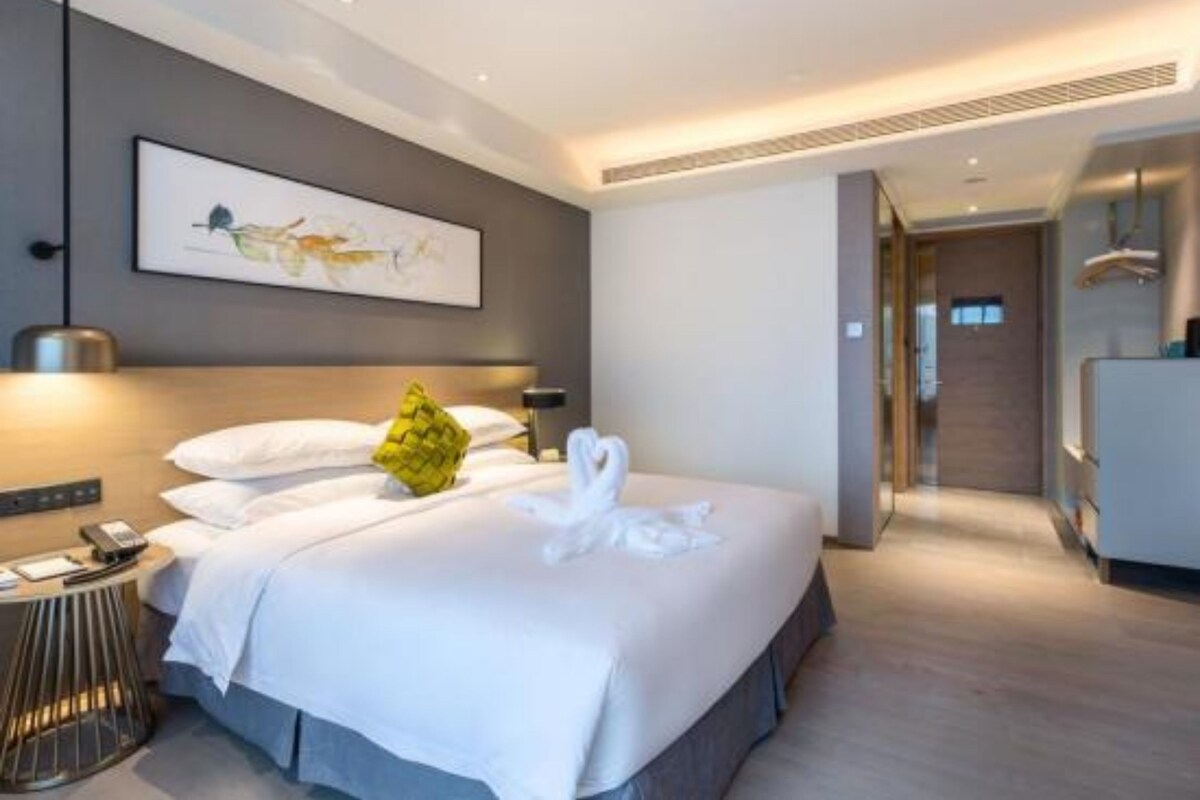 Une chambre d’hôtel moderne avec un grand lit, une table de chevet avec une lampe, une œuvre d’art encadrée au mur et une vue sur le couloir.