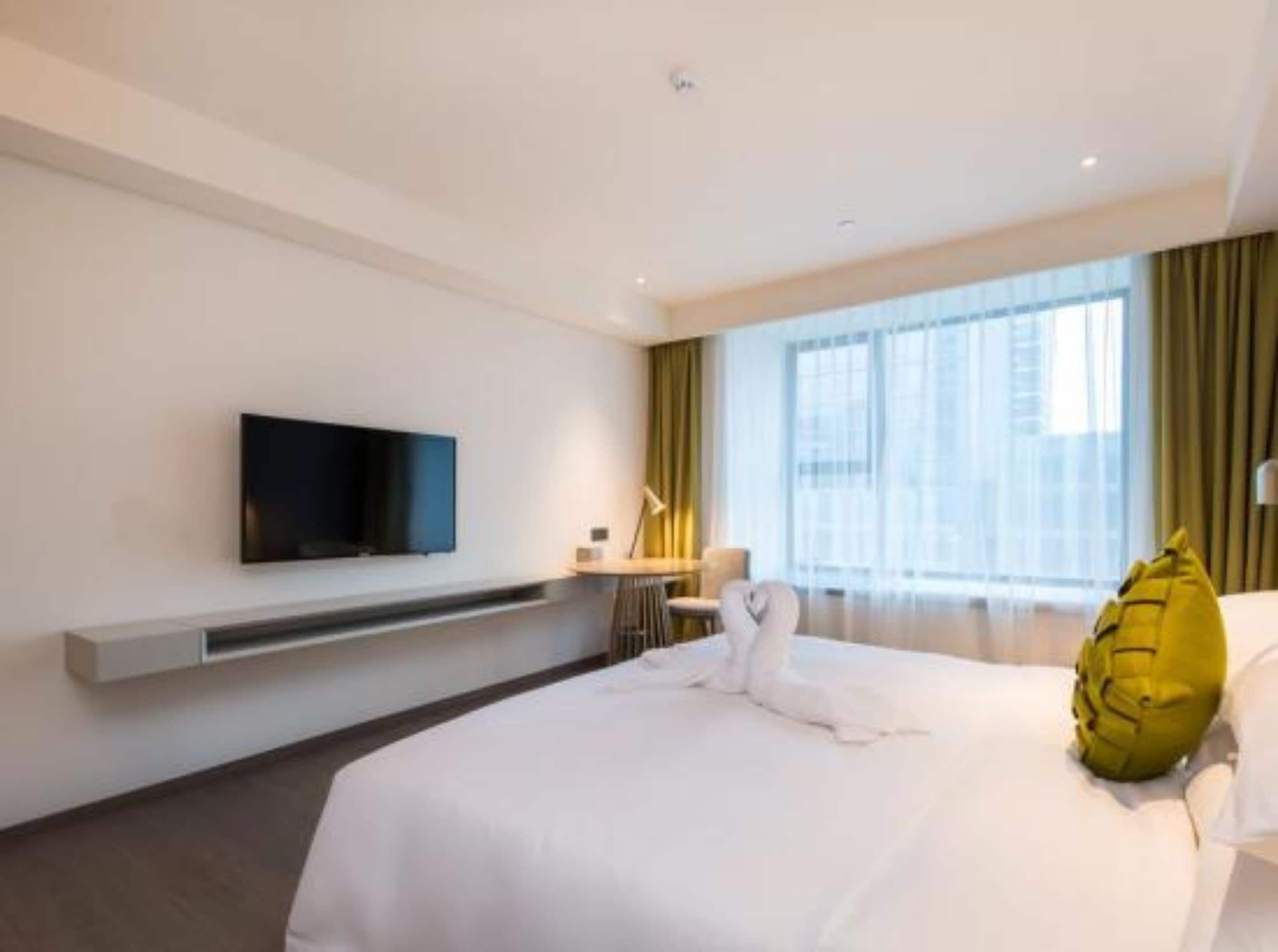 Una habitació d'hotel moderna amb un llit gran, una televisió de pantalla plana penjada a la paret, un escriptori amb una làmpada i una finestra ampla amb cortines.