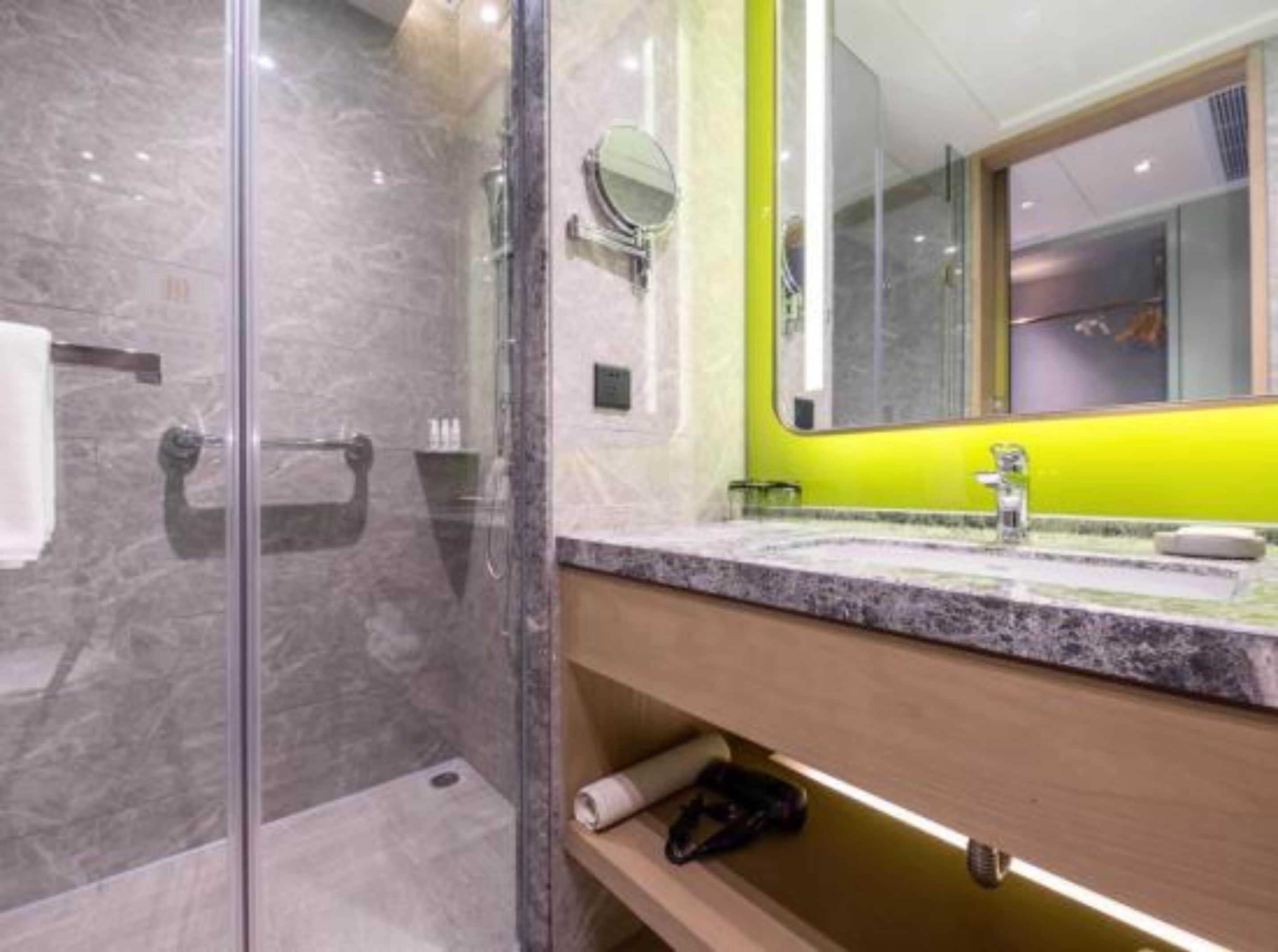 A modern bathroom with a glass shower enclosure, a marble-tiled floor, a wooden vanity with a sink, and a mirror with a green accent wall.