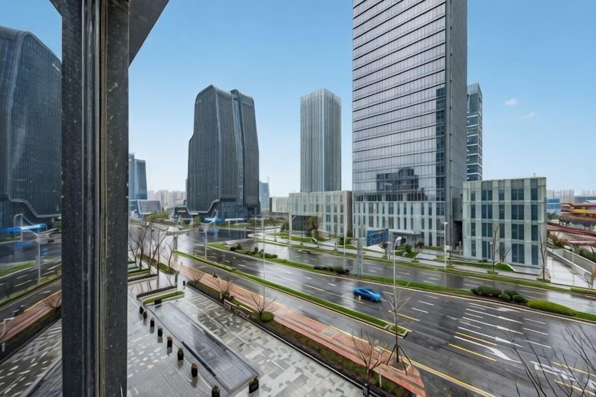A modern urban landscape with high-rise buildings, a wide road, and a pedestrian walkway.