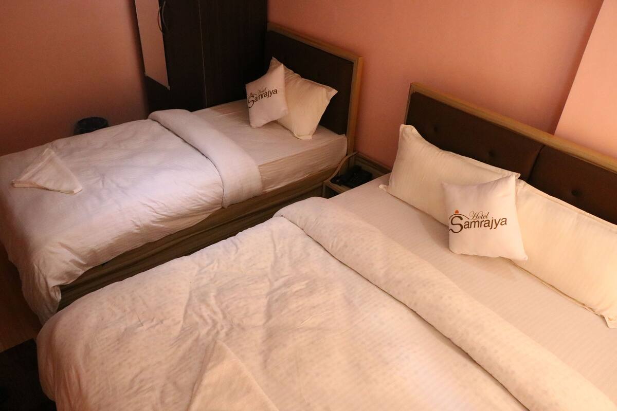 Dos camas individuales con ropa blanca de cama y almohadas, cada una con un cojín que muestra el logo del Hotel Samrajya.