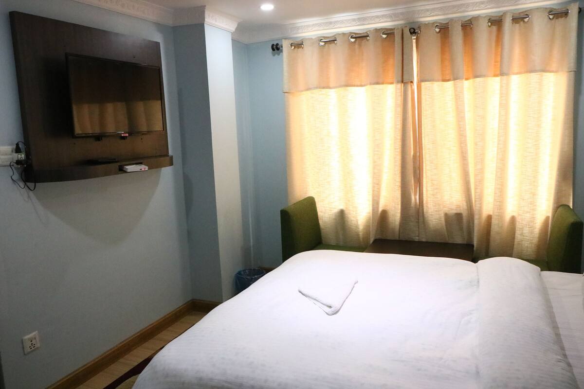 Habitación de hotel con cama, televisor de pantalla plana montado en la pared, ventana con cortinas y dos sillones verdes.
