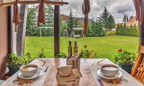 Une table avec deux bols de soupe, une assiette de pain et un récipient en métal contenant des couverts, le tout placé devant une fenêtre donnant sur un jardin avec une balançoire en bois et des arbres.