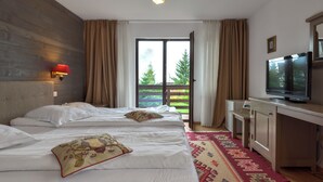 Ein Hotelzimmer mit Bett, Schreibtisch mit Fernseher, Fenster mit Vorhängen und Blick auf Bäume.