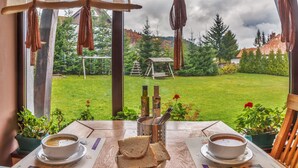 Une table avec deux bols de soupe, une assiette de pain et un récipient en métal contenant des couverts, le tout placé devant une fenêtre donnant sur un jardin avec une balançoire en bois et des arbres.