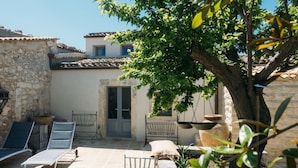 A courtyard with stone walls, a door, and a tree providing shade. There are lounge chairs and a small table.