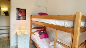 A bunk bed with white bedding and two pillows, a bedside table with a lamp, and a small white dresser with drawers.