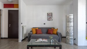 A living room with a grey sofa, red and yellow cushions, a glass coffee table with a book and remote control, and a white radiator. - Comfort House 14 (Roseto degli Abruzzi)