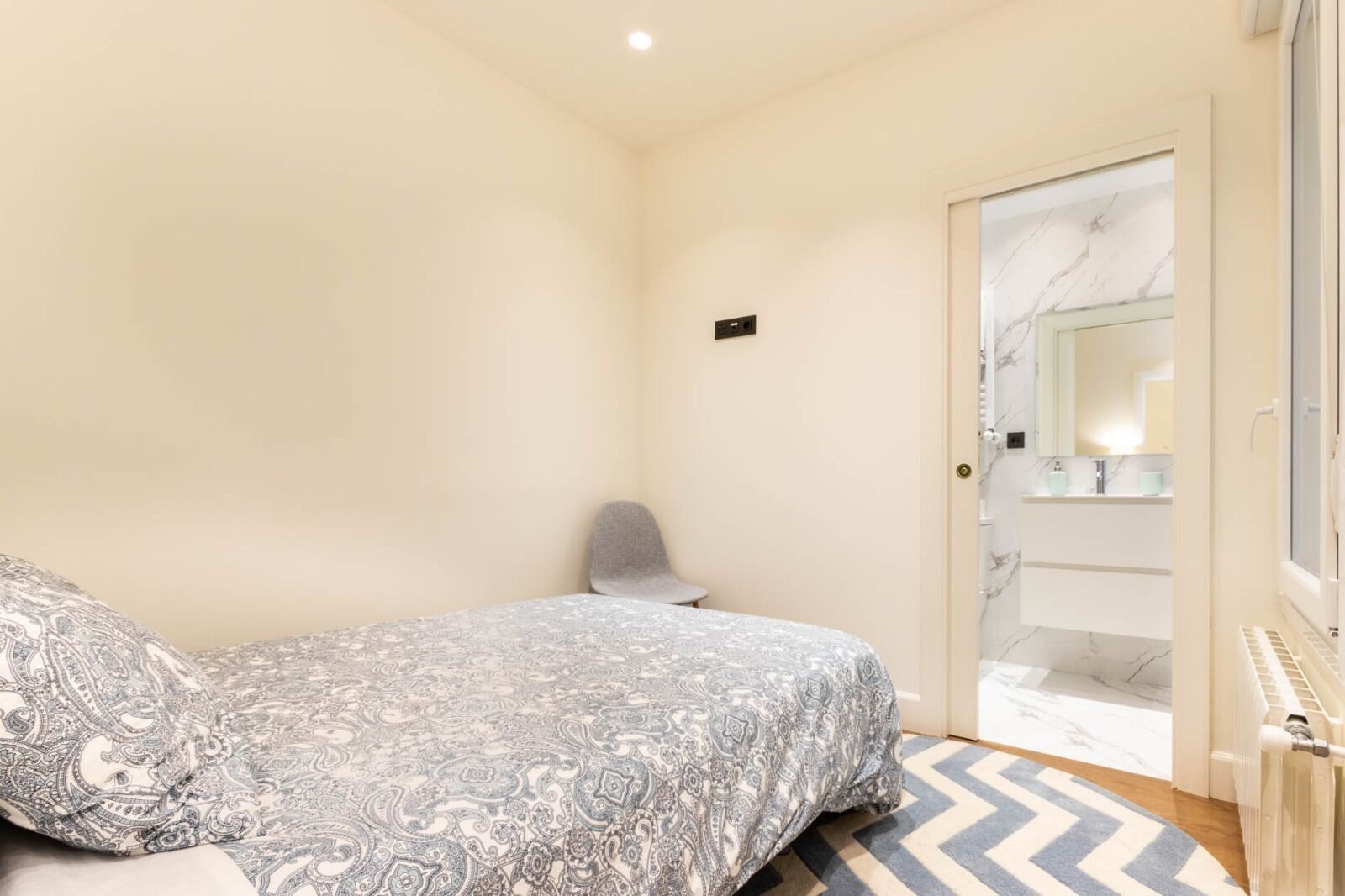 Un dormitorio con cama, una silla, una puerta que da a un baño y una alfombra estampada.