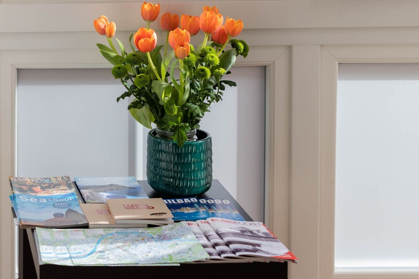 A table with a teal vase containing orange tulips, a map, and several travel magazines.