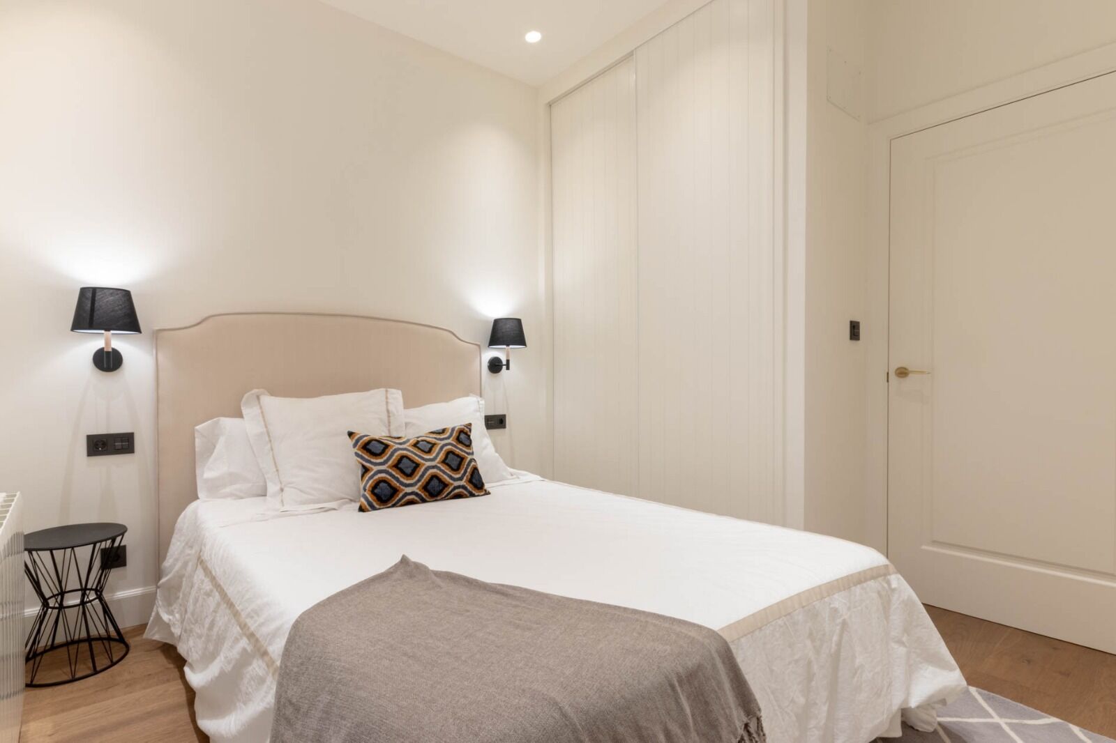 Un dormitorio ordenado con cama, dos lámparas de noche, una mesita de noche y una puerta.