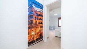 Corredor com um mural vibrante retratando uma paisagem urbana, parede branca e área de estar com sofá branco e cadeiras de plástico transparente.