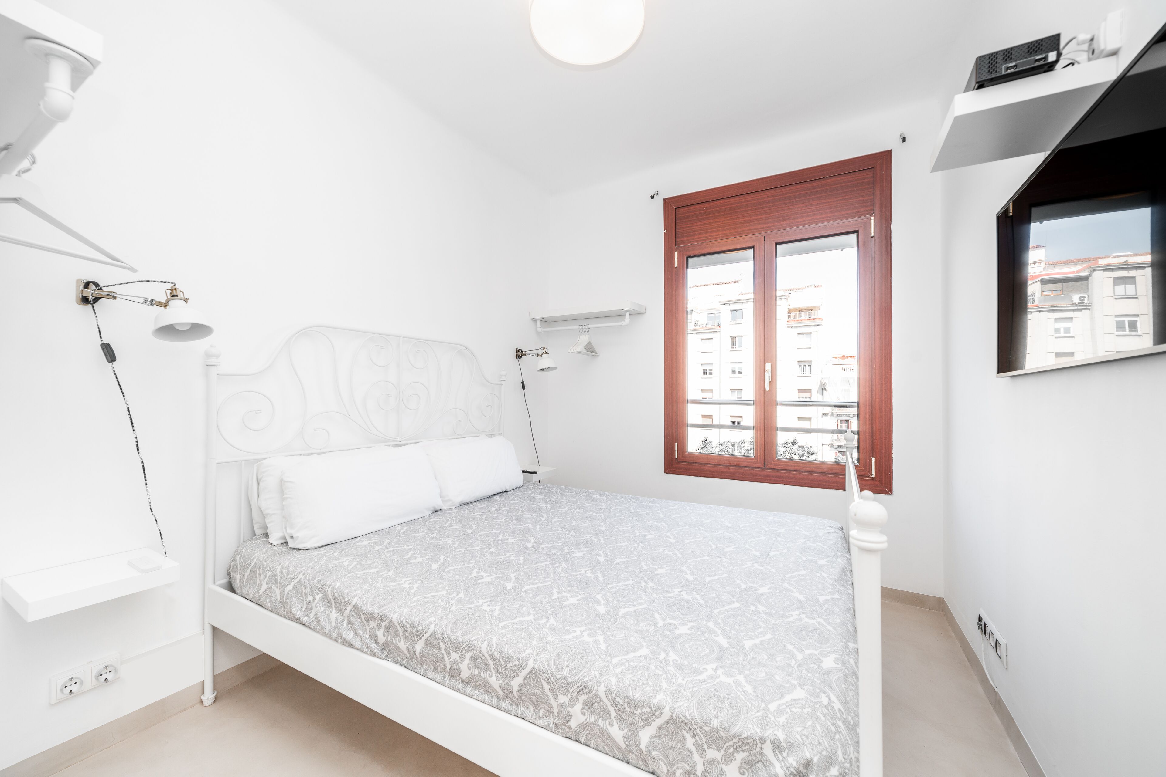 Ein Schlafzimmer mit einem Bett, einem Nachttisch, einer Lampe, einem Fenster mit Blick auf Gebäude und einem wandmontierten Regal.