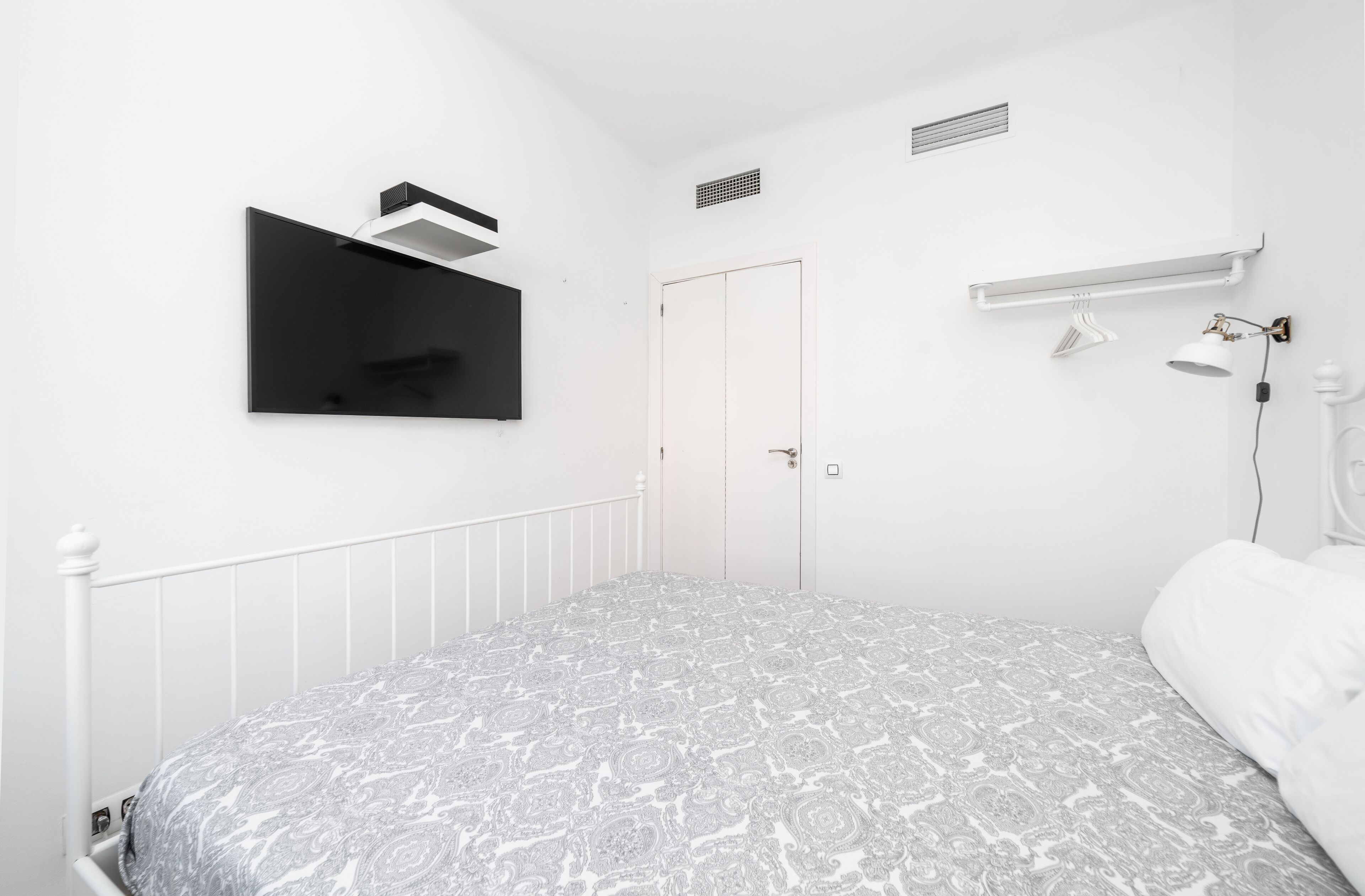 A bedroom with a bed, a wall-mounted TV, a door, a white radiator, and a hanging lamp.