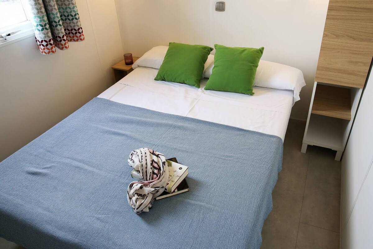 Un lit bien fait, recouvert d’une couvre-lit bleue et agrémenté de deux coussins verts, une petite table de chevet avec une bougie, et une étagère en bois sur le côté droit.