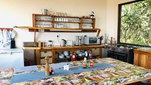 A kitchen with a wooden table covered in a colorful tablecloth, a microwave, and various kitchen utensils. There is a window with a view of trees. - Topo Das Arvores (Ponta d'Ouro)