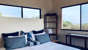 A bedroom with a large bed, a nightstand, a lamp, and a bookshelf. There are two windows providing a view of the outdoors. - Topo Das Arvores (Ponta d'Ouro)