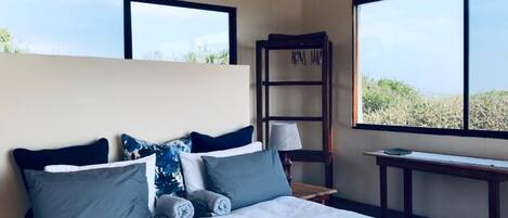 A bedroom with a large bed, a nightstand, a lamp, and a bookshelf. There are two windows providing a view of the outdoors.
