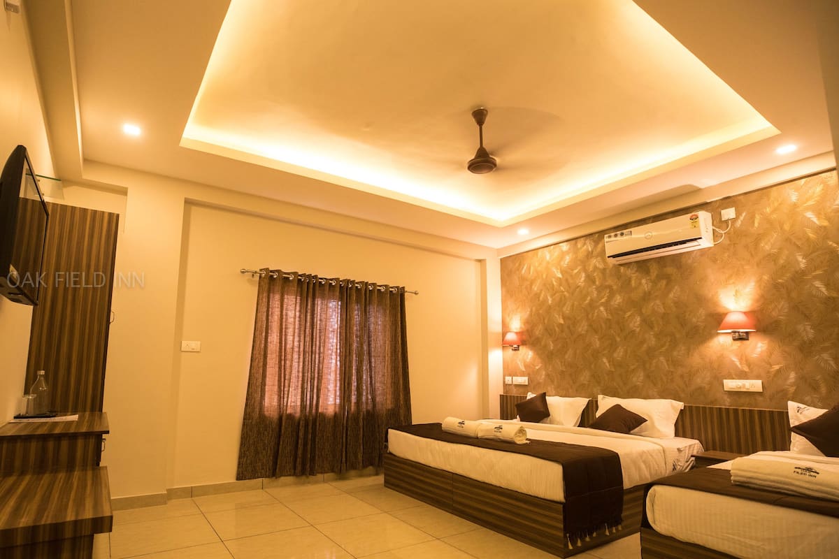 A hotel room with a bed, a desk, a mirror, a window with curtains, an air conditioning unit, and wall-mounted lights.